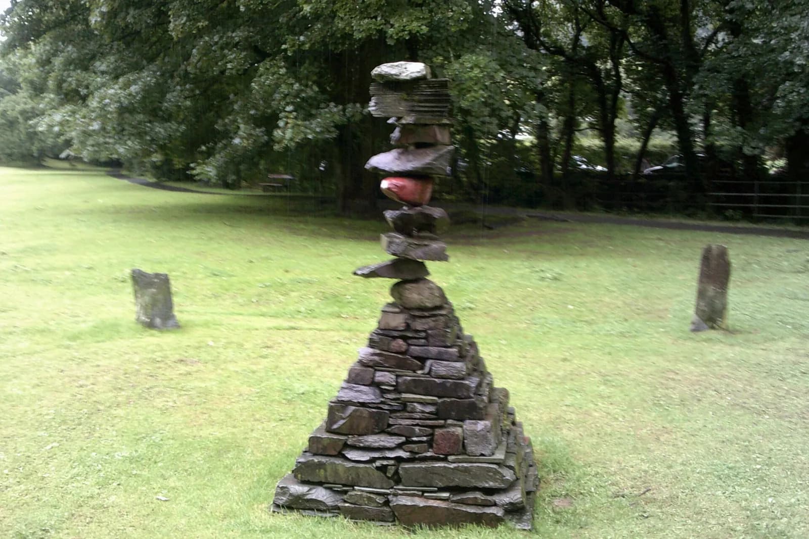 A stacked-stone sculpture on the grass of Rothay Park in Ambleside, with trees behind