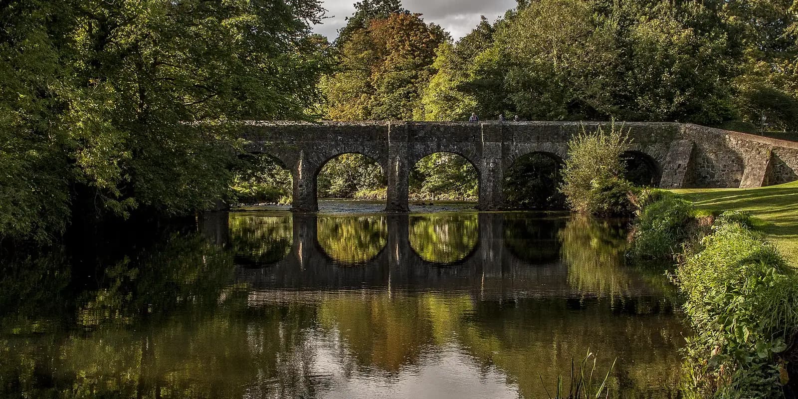 The old stone Deerpark Bridge reflected in still water among trees, in the grounds of Antrim Castle