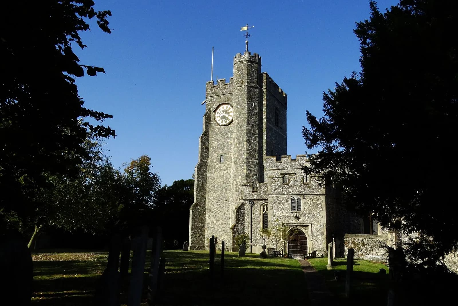 The flint tower of St Mary's Church in Chilham, framed by dark trees against a blue sky