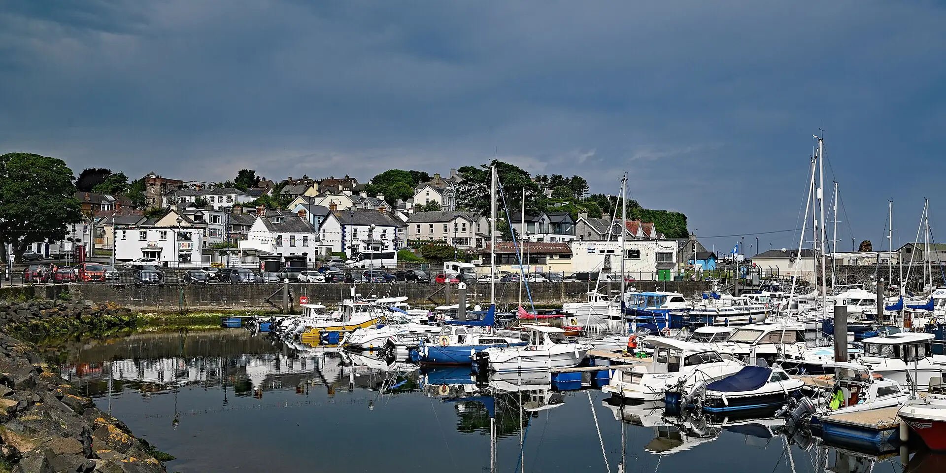 Boats moored in Ballycastle marina, with white houses along the shore under a stormy sky