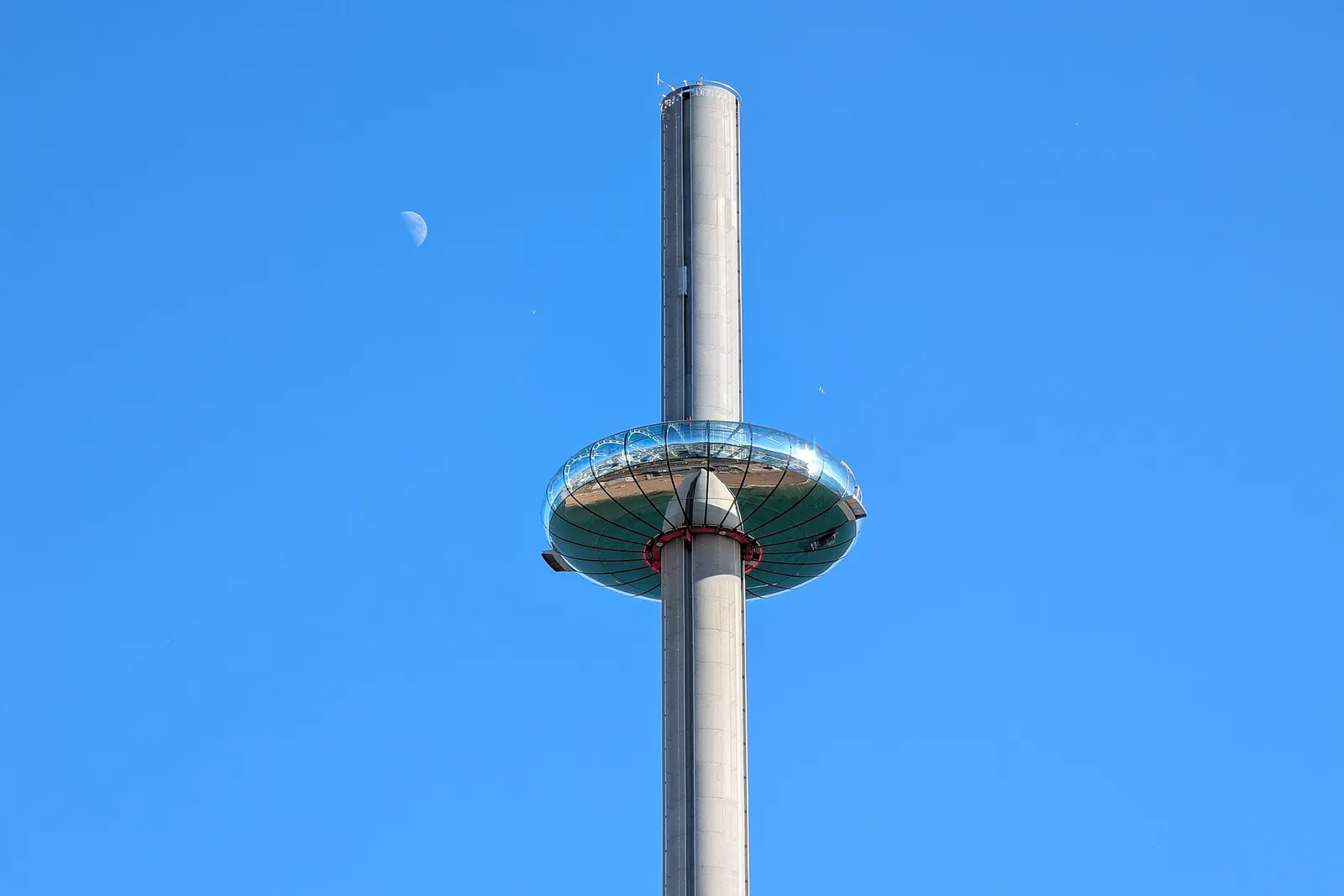 The slim tower and glass pod of the Brighton i360 against a clear blue sky, with the moon above