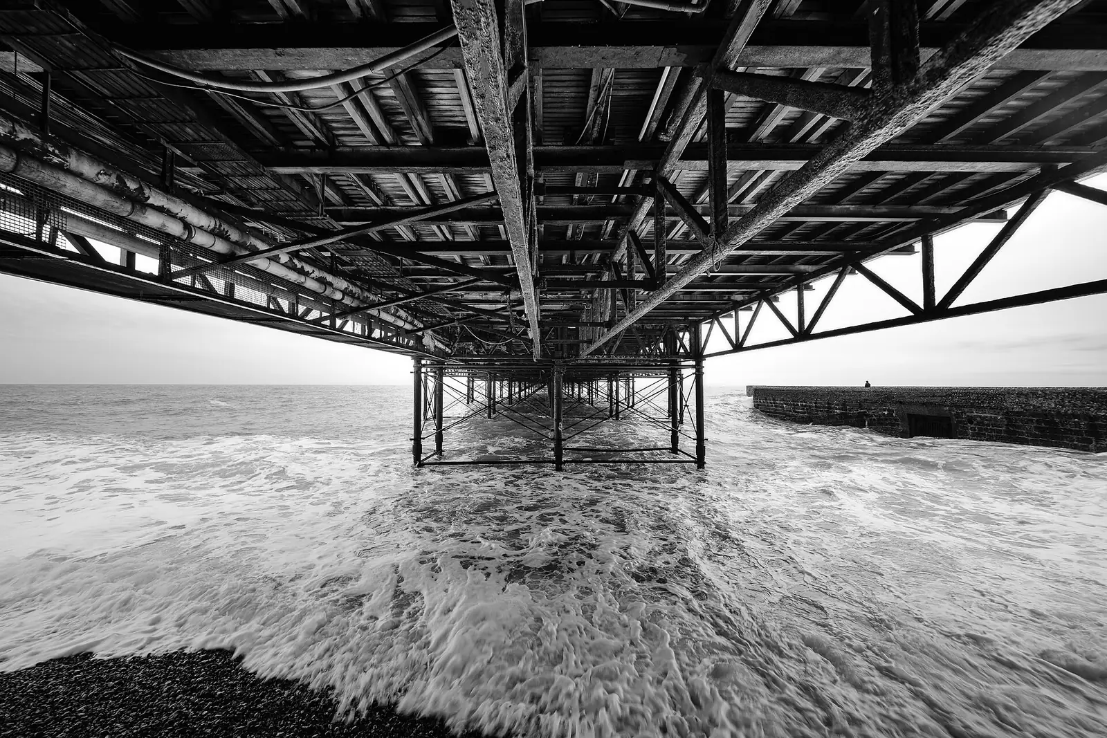 Waves rushing in beneath the iron legs of Brighton Palace Pier, in black and white