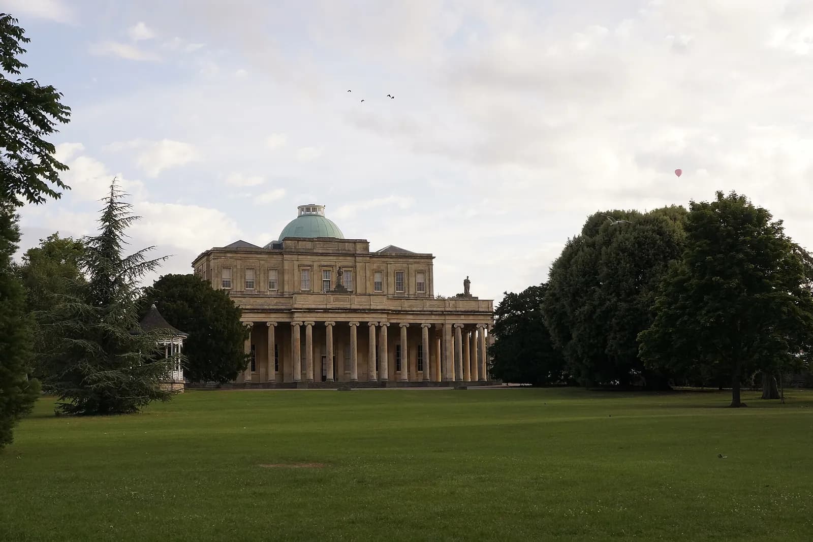 The domed and columned Pittville Pump Room beyond a wide lawn and trees
