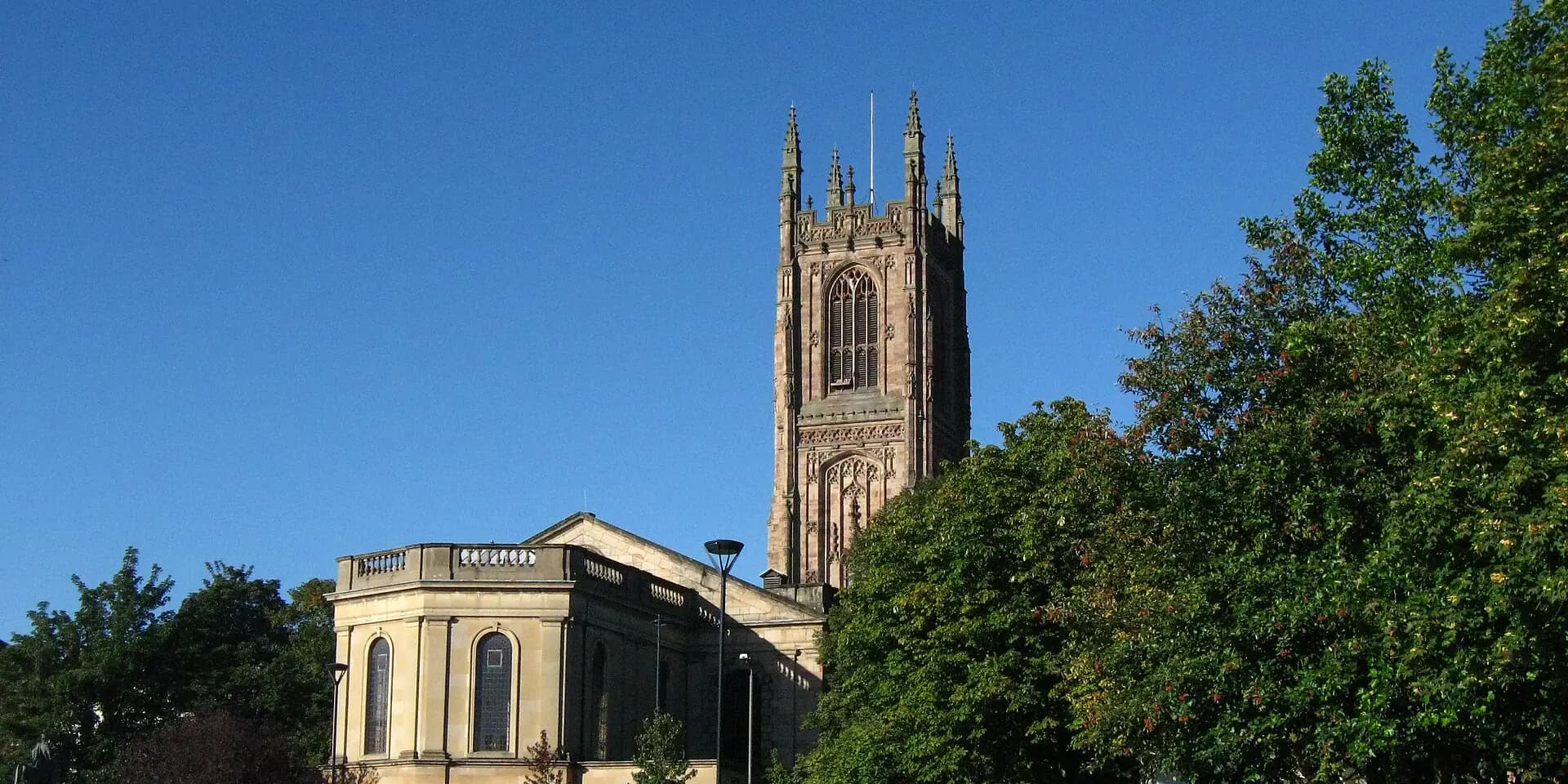 The tall pinnacled tower of Derby Cathedral among trees under a blue sky