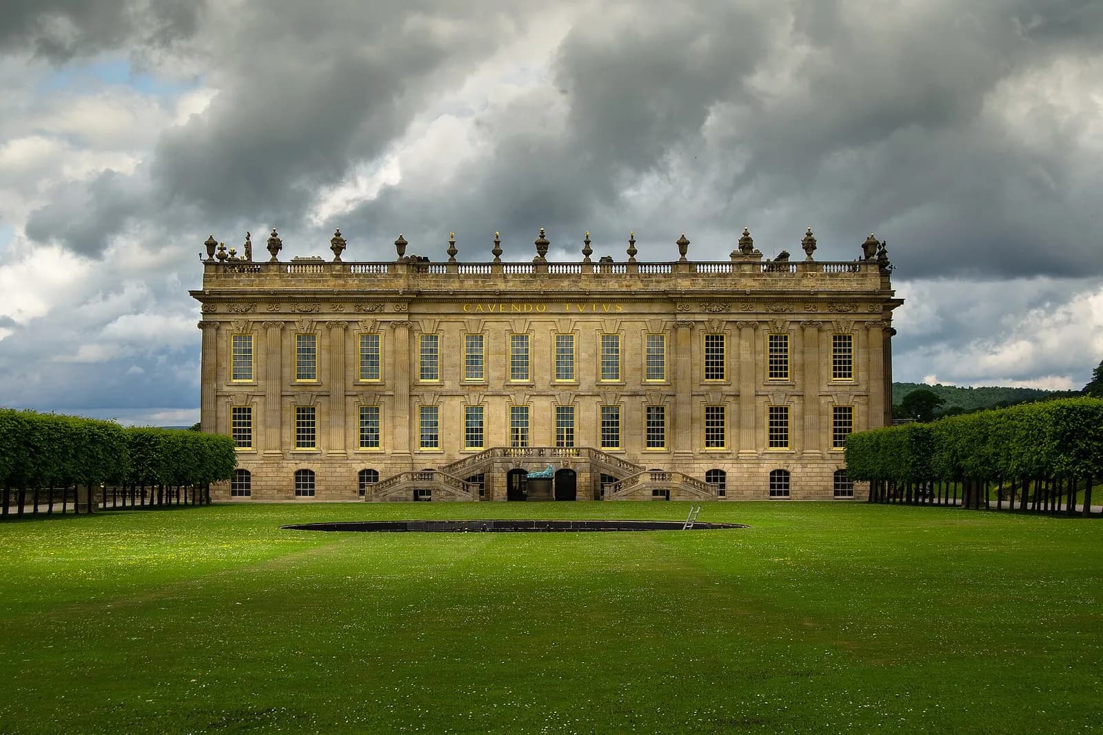 The golden south front of Chatsworth House above a wide lawn under a stormy sky