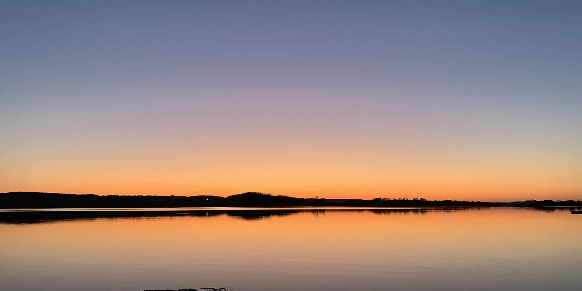 Sunset over still water on Lower Lough Erne, with a low dark shoreline on the horizon