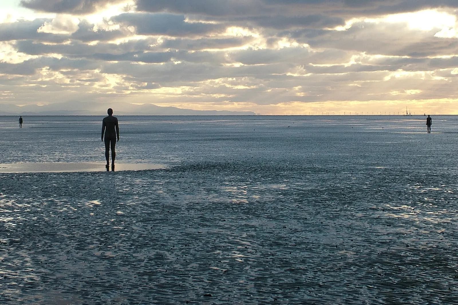 One of Antony Gormley's iron figures standing on Crosby beach, looking out to sea under a cloudy sky