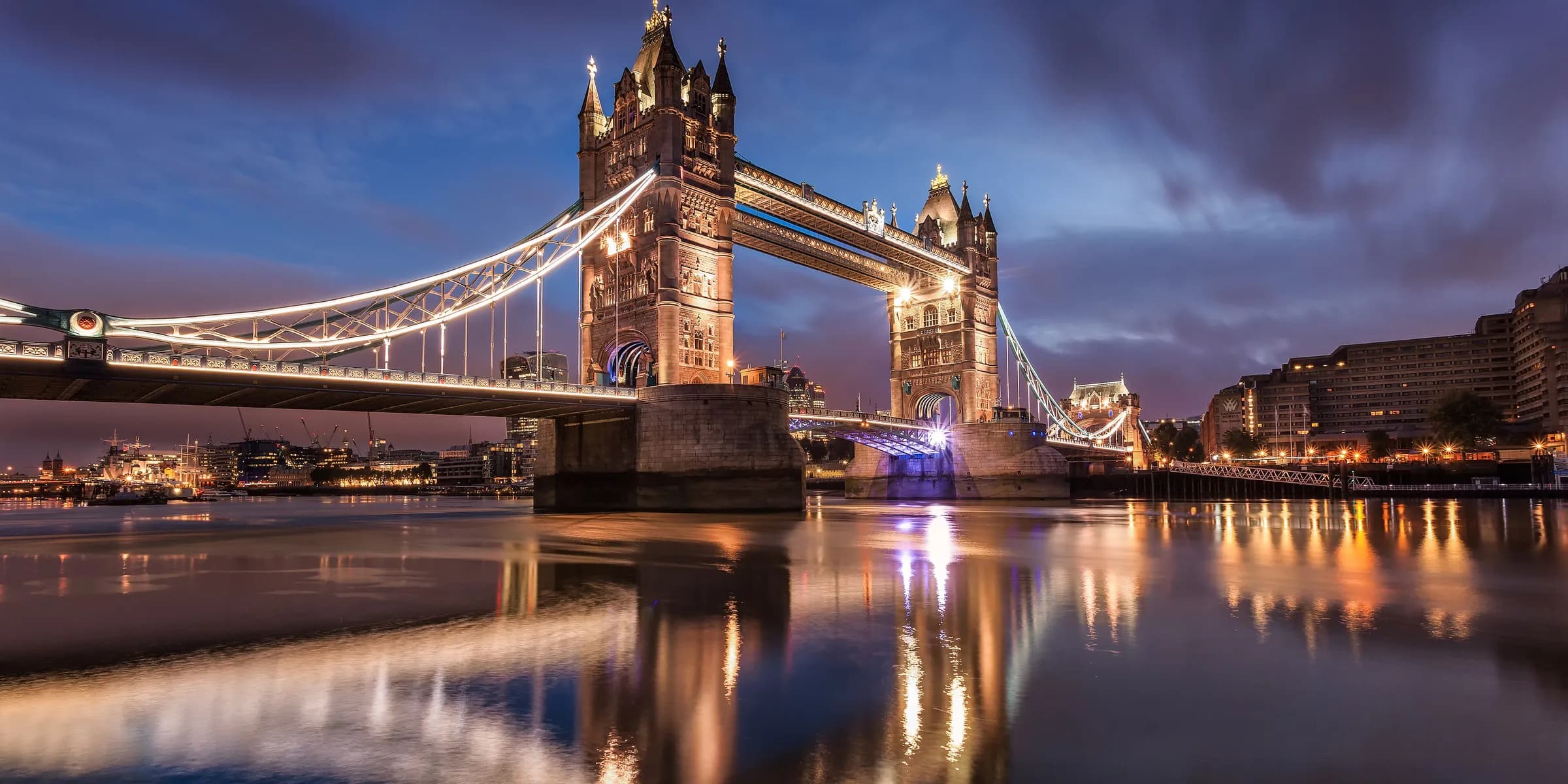 Tower Bridge lit up at dawn, its towers reflected in the Thames under a purple sky
