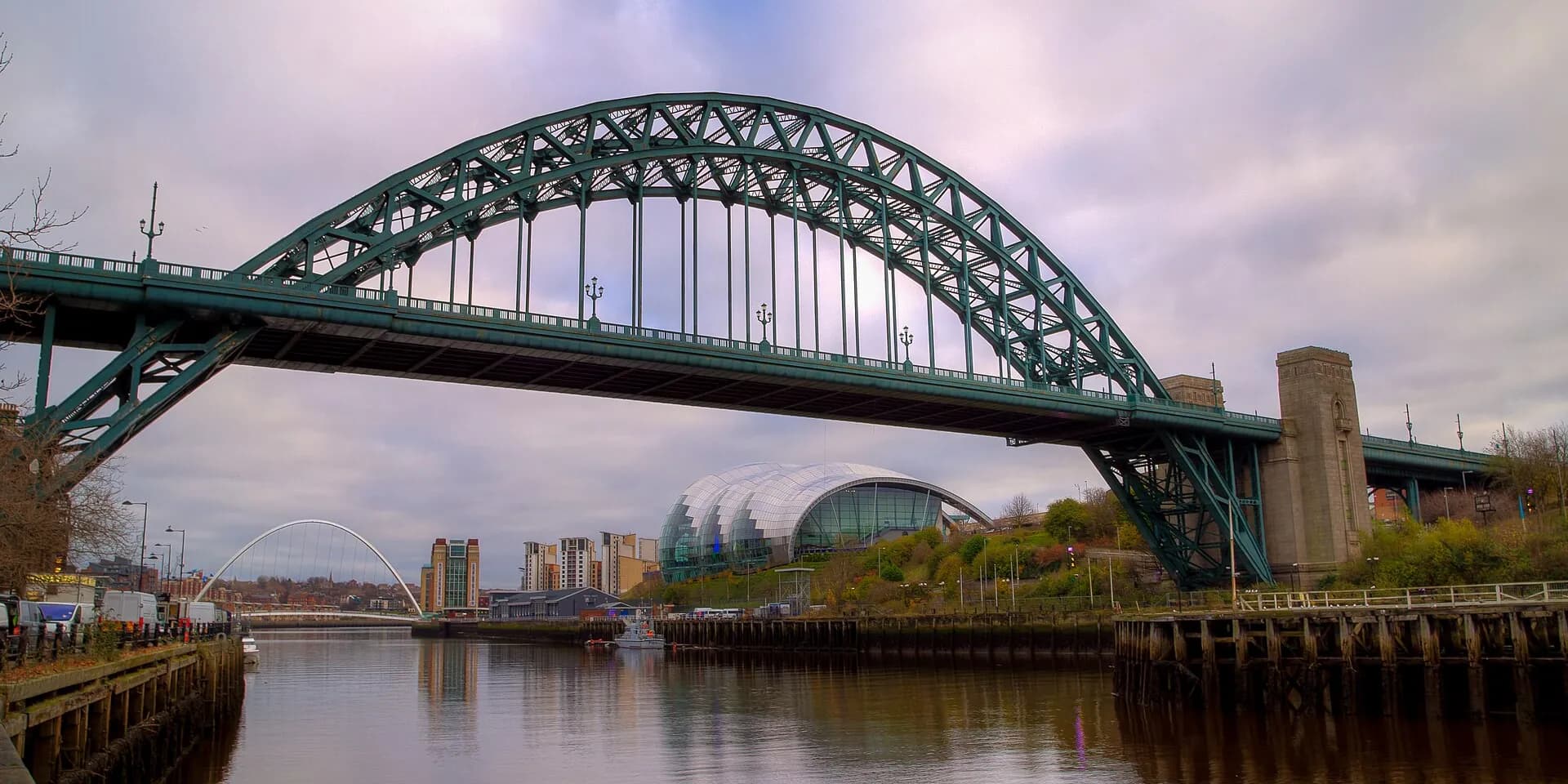 The green steel arch of the Tyne Bridge over the river, with the Sage building on the Gateshead bank