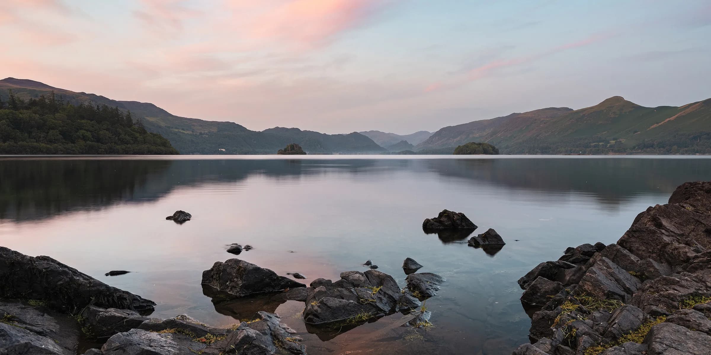 Still water on Derwentwater at first light, with rocks in the foreground and fells under a pink sky