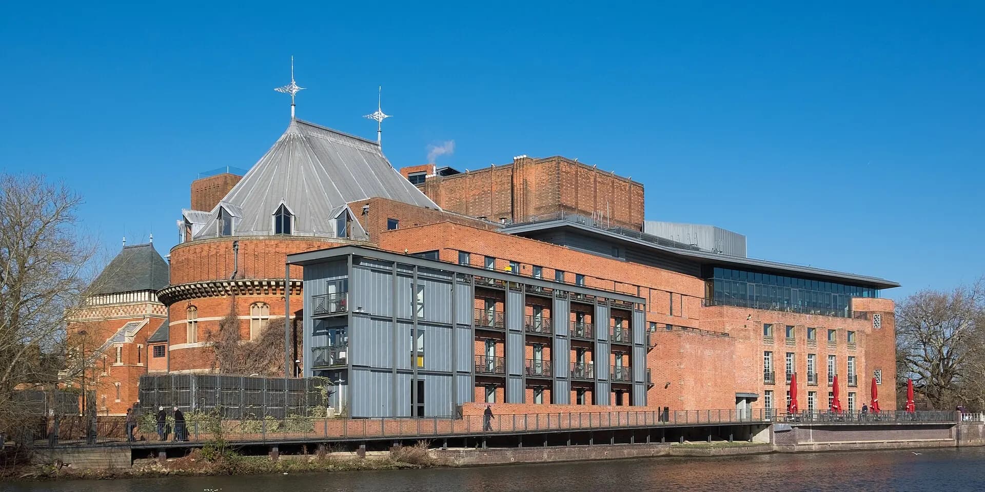 The red-brick Royal Shakespeare Theatre beside the River Avon, under a blue sky