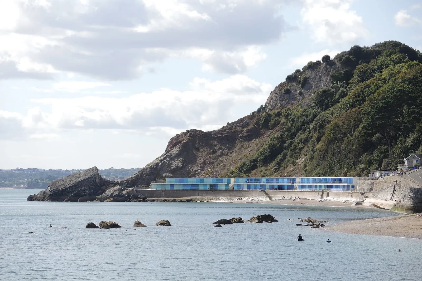 Meadfoot beach below a steep wooded headland, with swimmers in the calm sea