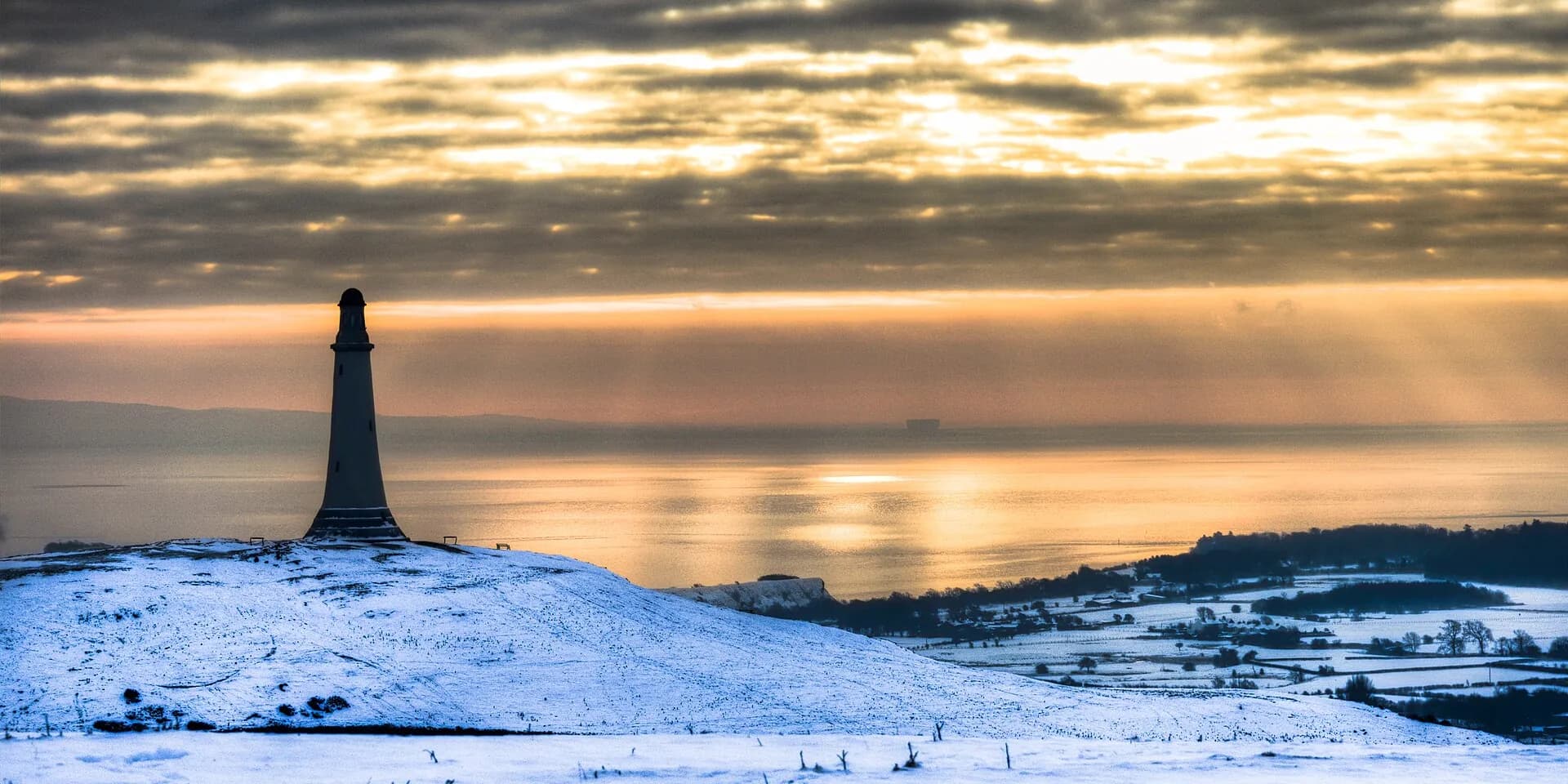 The Hoad monument, a lighthouse-shaped tower on a snowy hill above Morecambe Bay at sunset