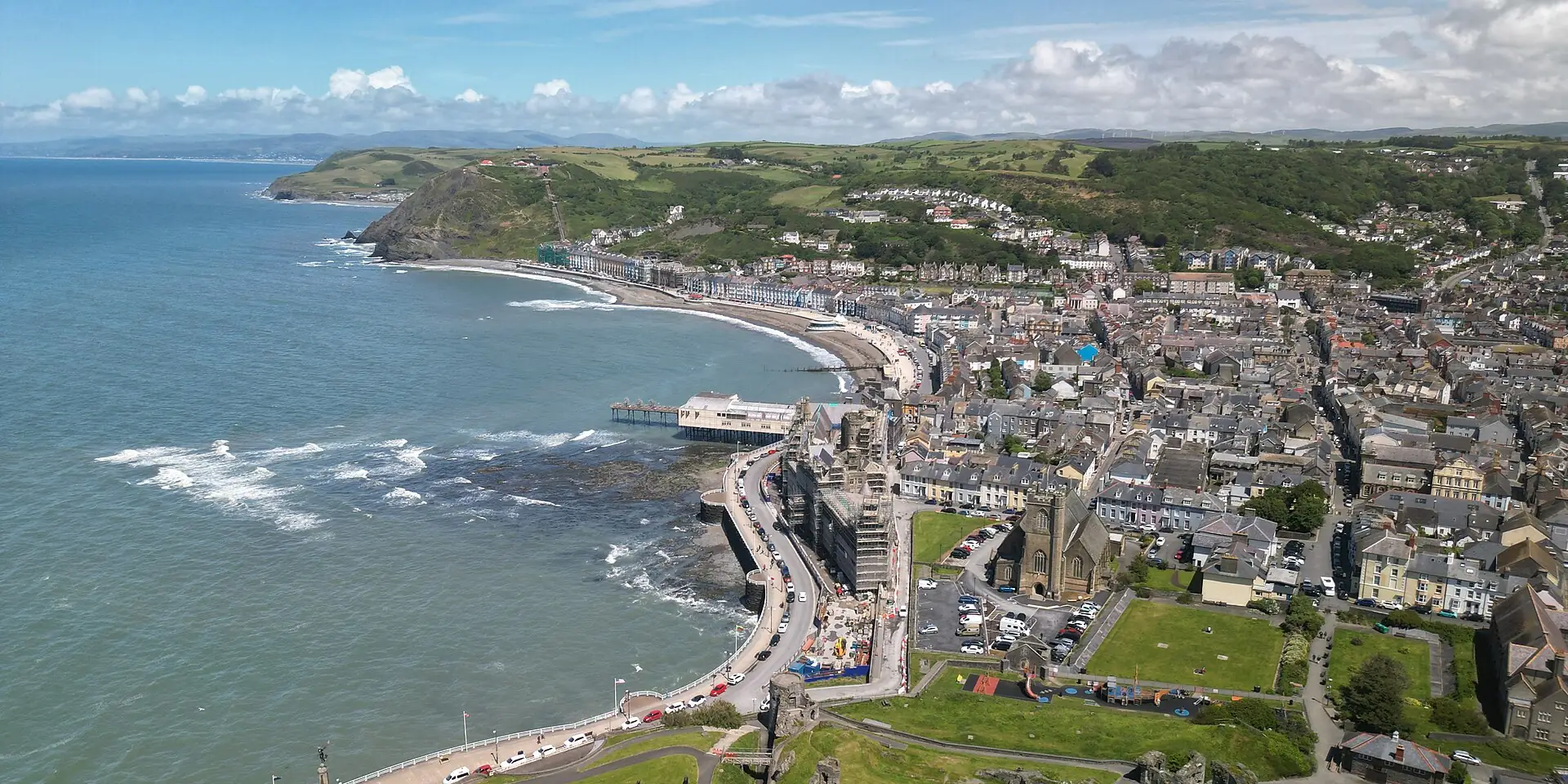 Aberystwyth from the air: the curving seafront and pier, the town's rooftops and green hills beyond