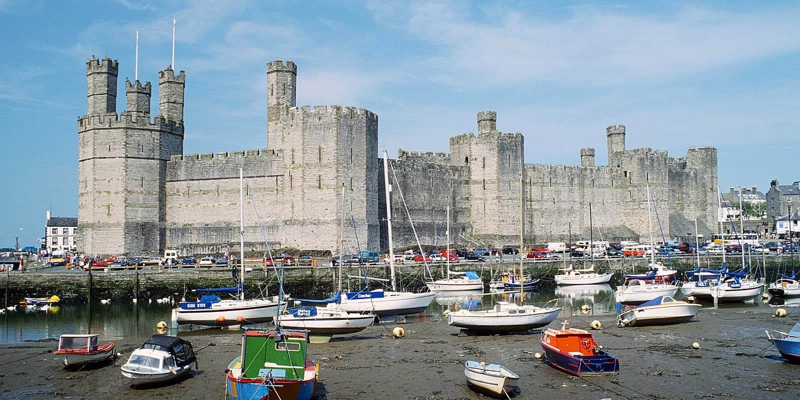 Caernarfon Castle's great stone towers above the harbour, with small boats at low tide