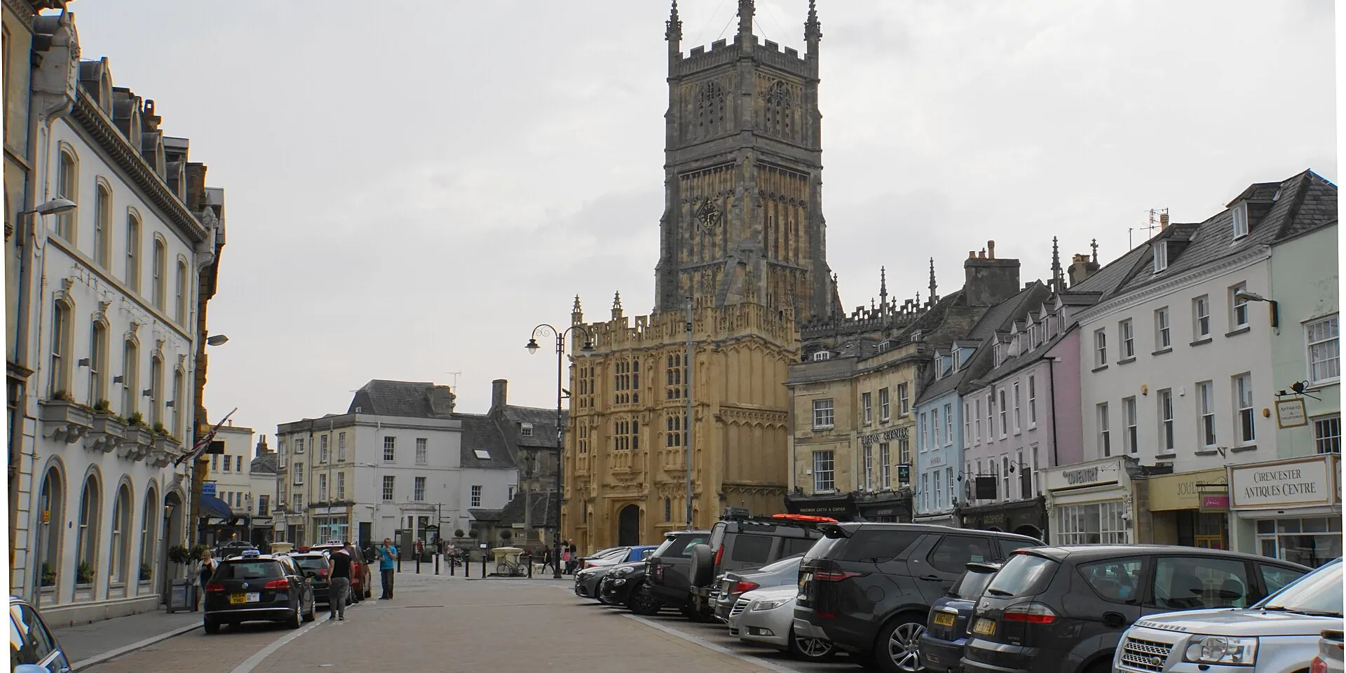 Cirencester's Market Place, lined with stone and painted buildings, with the parish church tower at the end