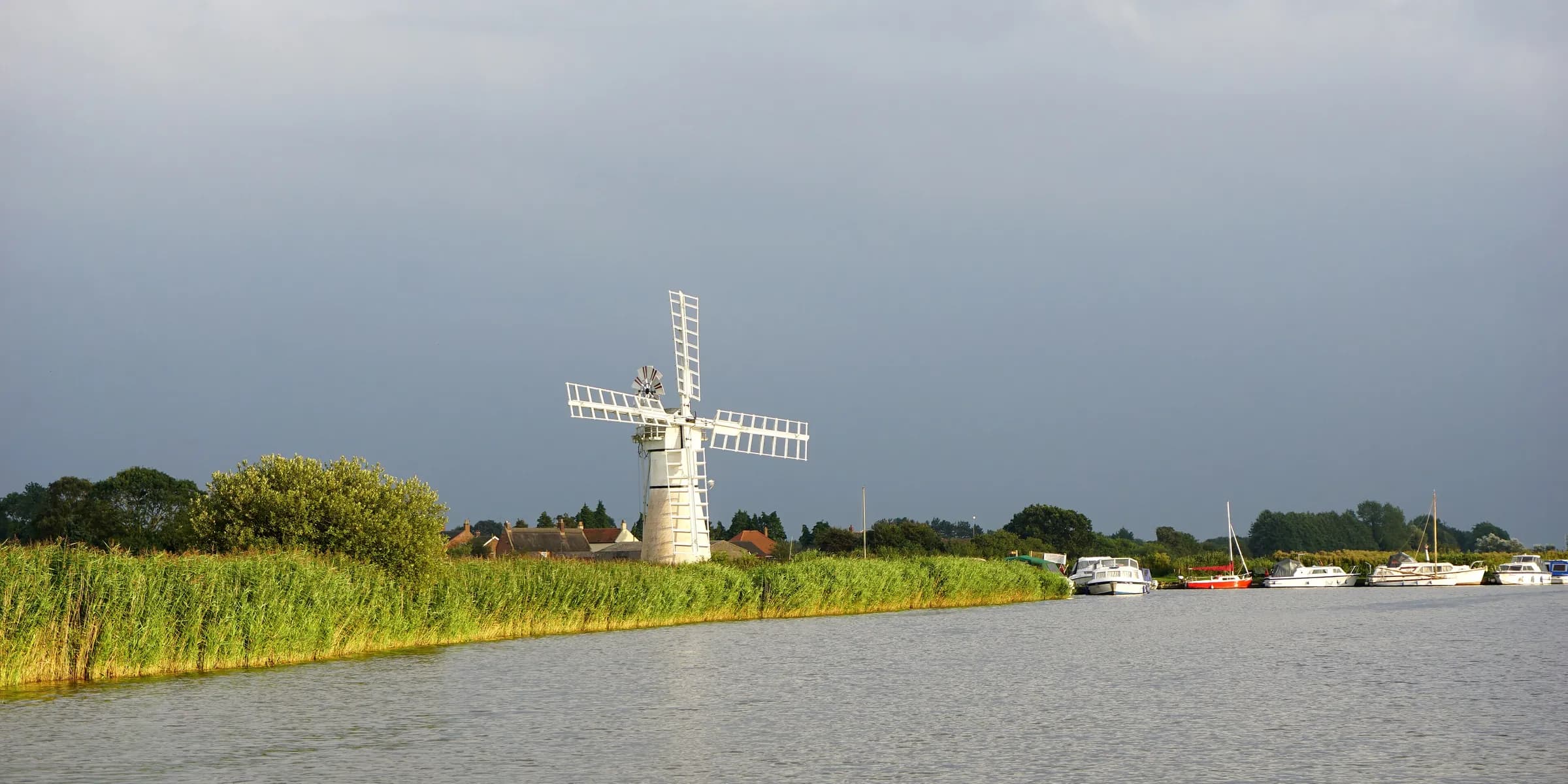 A white windpump beside the River Thurne, with reeds along the bank and boats moored further along