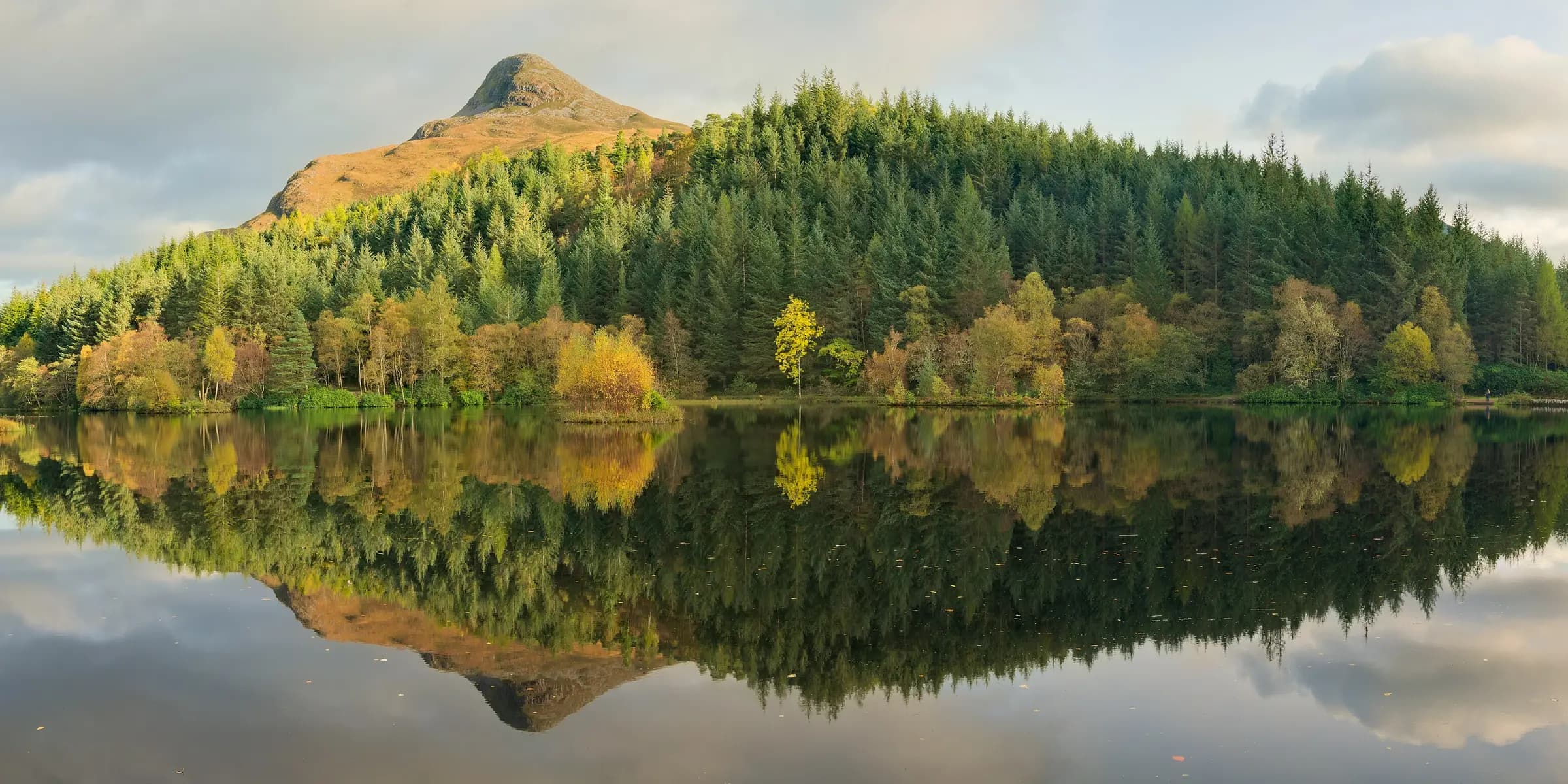 Pine and autumn-coloured trees reflected in a still lochan, with the pointed Pap of Glencoe behind