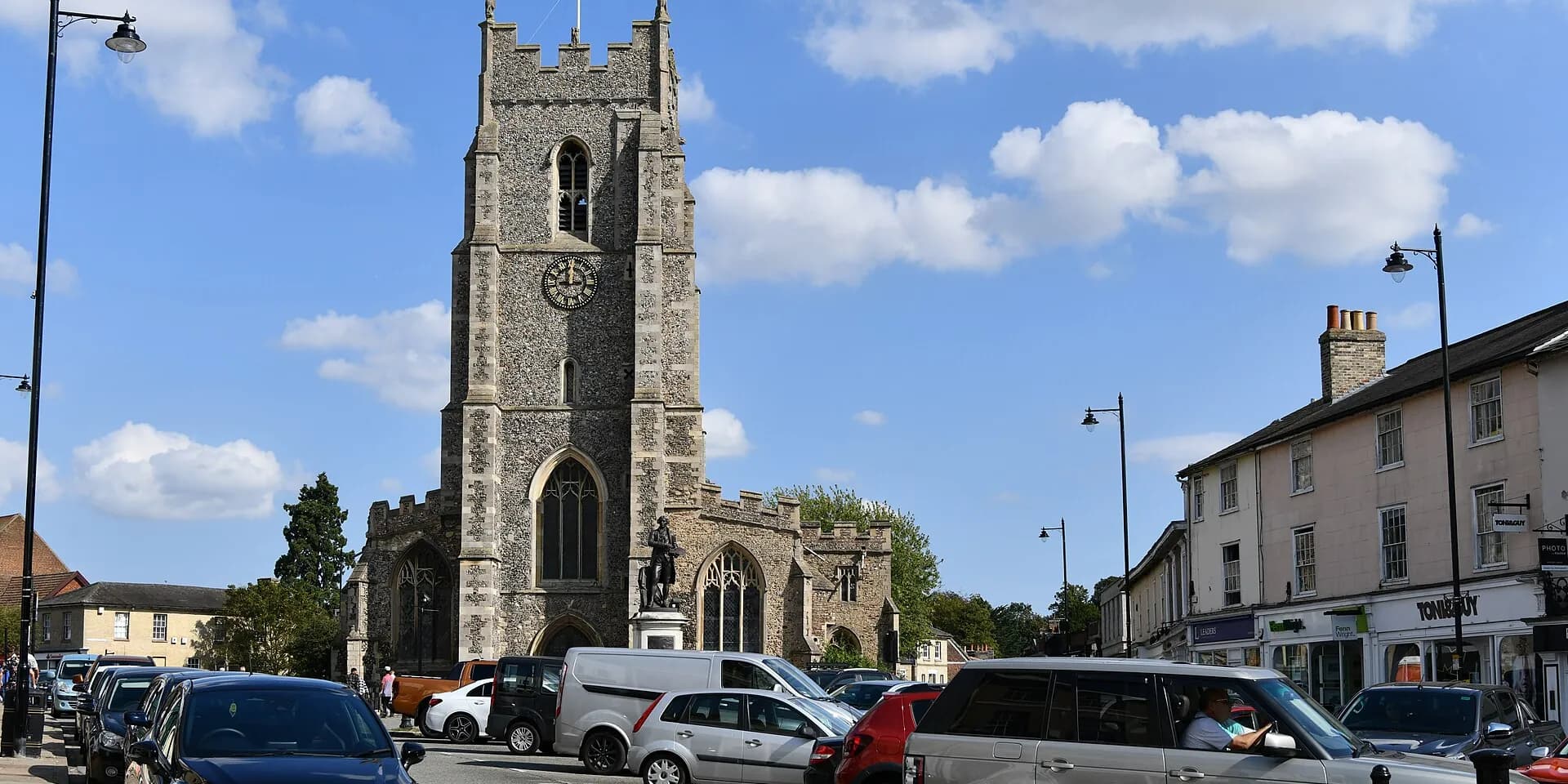 The flint tower of St Peter's Church on Market Hill in Sudbury, with Georgian buildings alongside under a blue sky