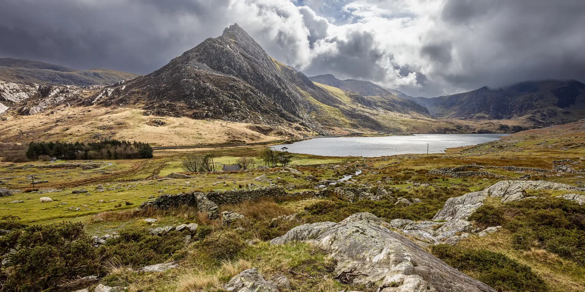 The rocky peak of Tryfan rising above Llyn Ogwen, with rough moorland and boulders in front under heavy cloud