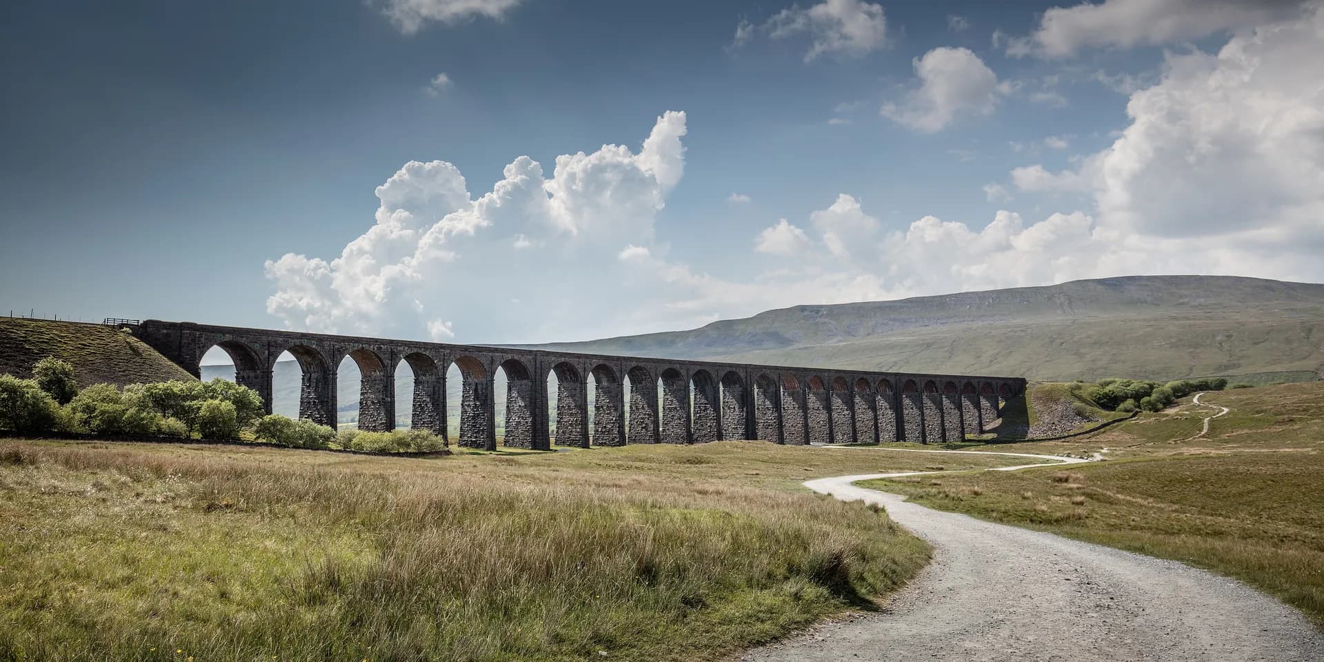 The stone arches of Ribblehead Viaduct crossing open moorland, with a gravel track winding towards it under a cloudy sky